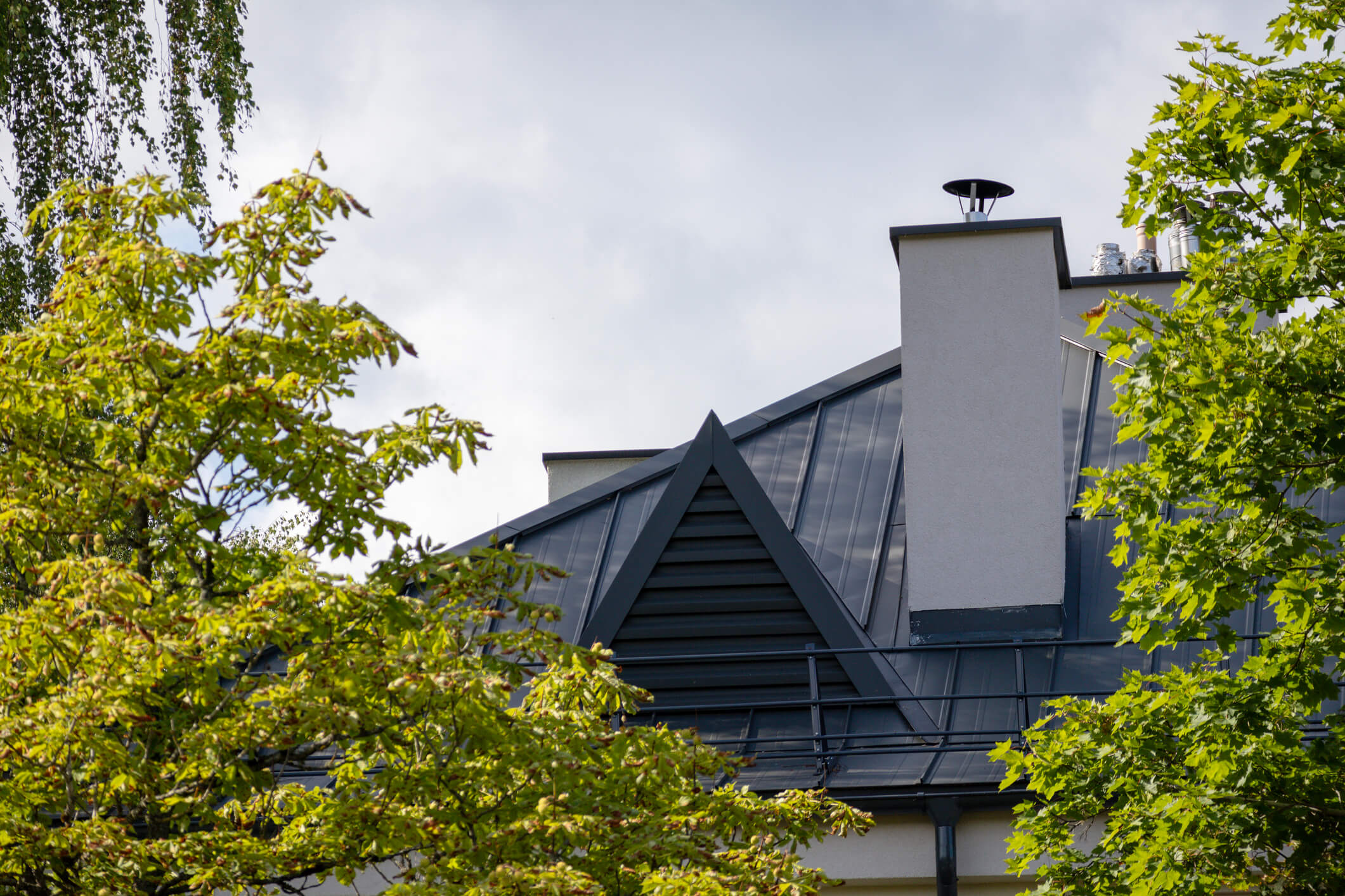 Modern house with dark metal roof and white chimney surrounded by green trees